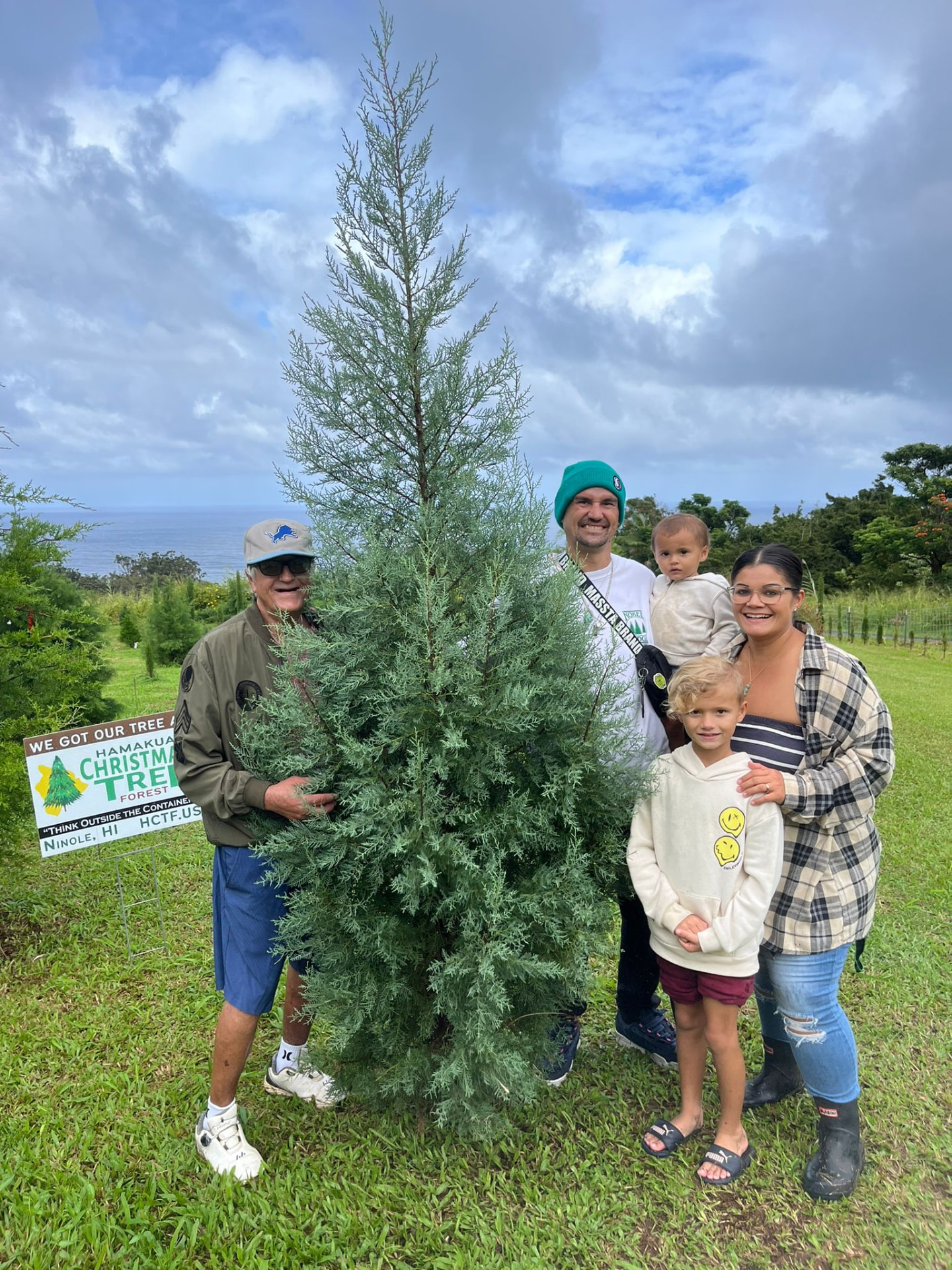 Family of five posing with their blue-green Christmas tree and a Hamakua Christmas Tree Forest sign